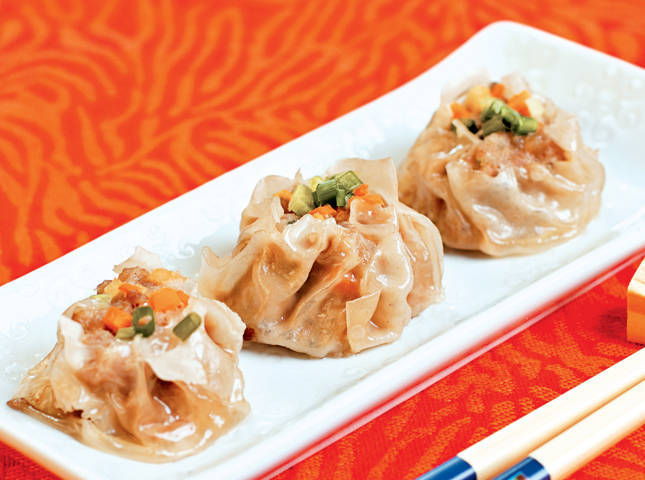 Three steamed siomai or chinese pork dumplings wrapped in thin wrappers, topped with carrots and spring onions on a white platter