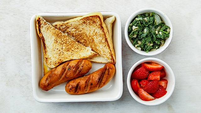 bread and sausage in a white square bowl with greens and strawberry in different bowls on the side