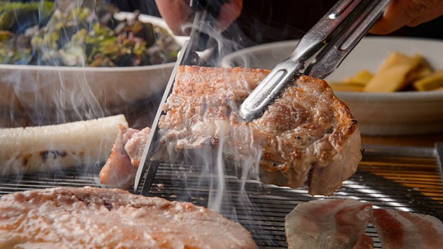 a piece of pork belly or samgyupsal being lifted from the grill with tongs