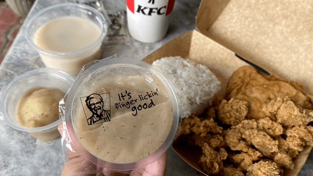KFC gravy being held up against a background with KFC chicken, hotshots, mashed potato, and mushroom soup